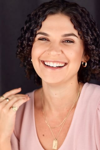 Woman with dark curly hair smiling at camera, wearing a pink button-up shirt and gold chain necklace with pendant against a dark background.